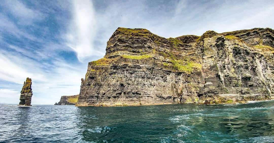 Scenic view of the Cliffs of Moher and a sea stack along the Irish coastline under a partly cloudy sky.