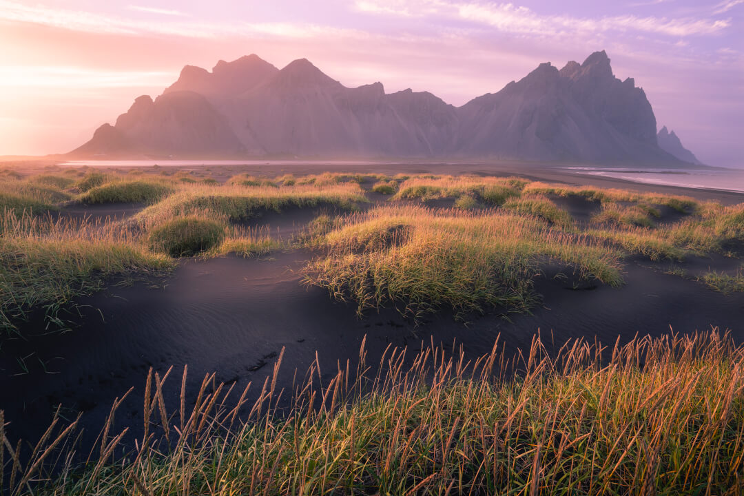 Serene mountain landscape with black sand beach and grass dunes under a pastel sky in Iceland.
