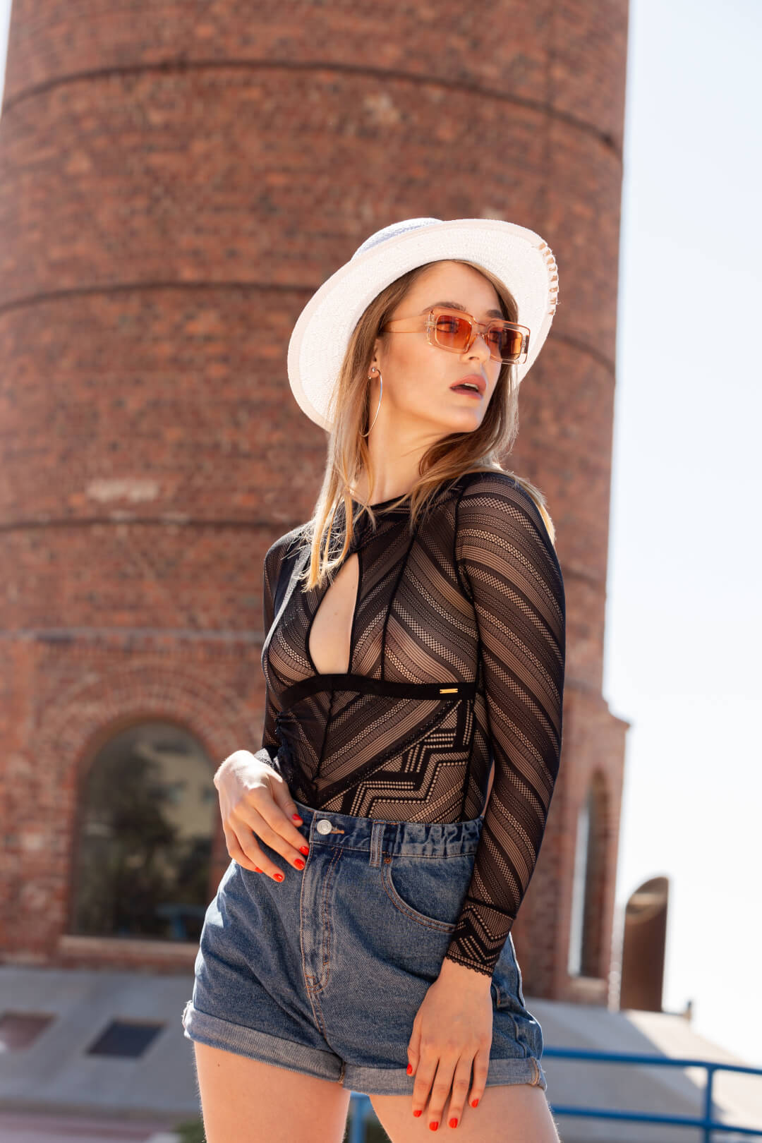 A stylish woman poses in a black bodysuit, denim shorts, hat, and sunglasses against an urban background.