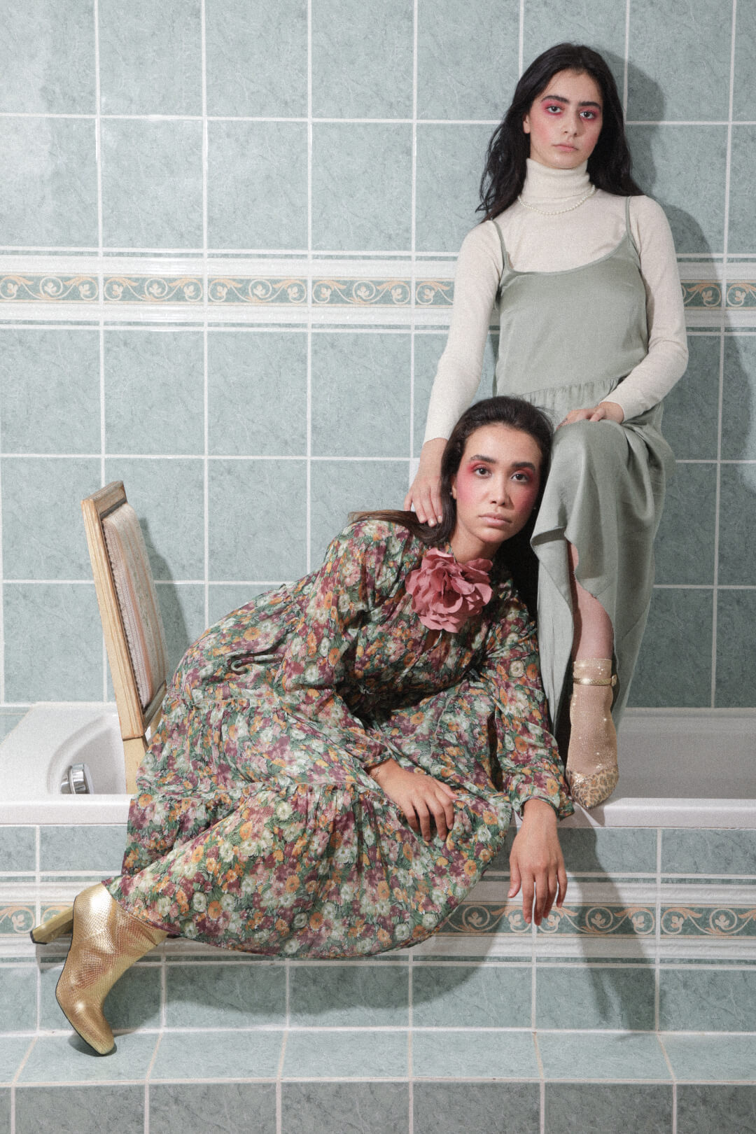 Two women pose in a bathroom setting, showcasing vintage-inspired fashion with soft, muted tones for an editorial look.