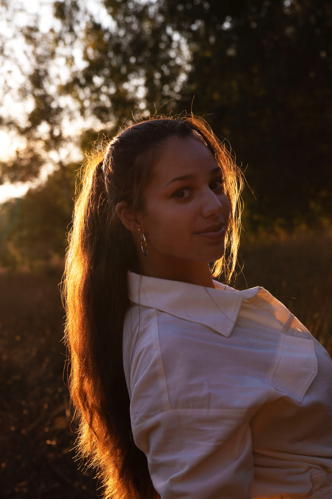 A young woman with long hair smiles as she turns to face the camera in a sunlit outdoor setting.
