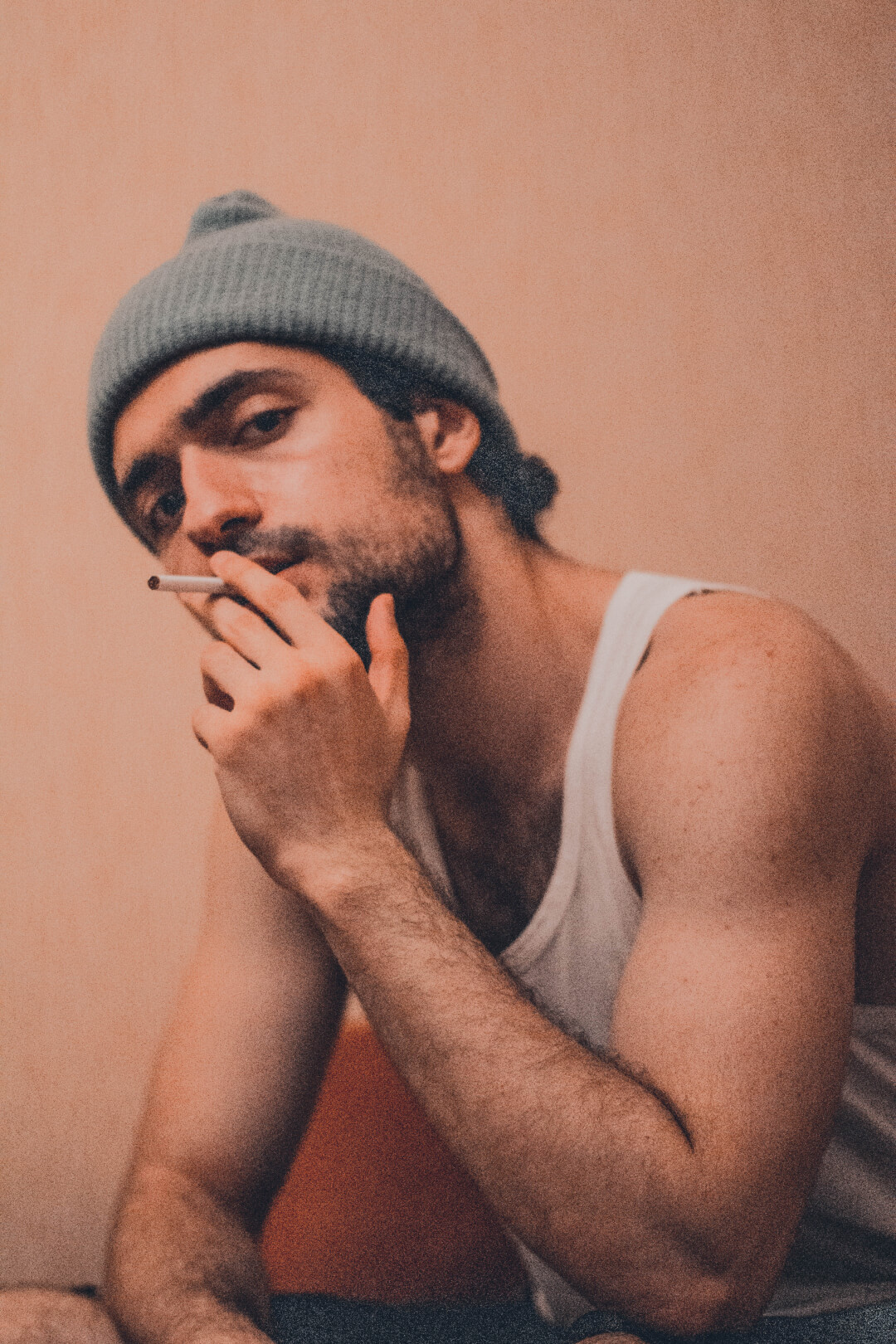 Portrait of a man with a beanie and tank top smoking a cigarette indoors.