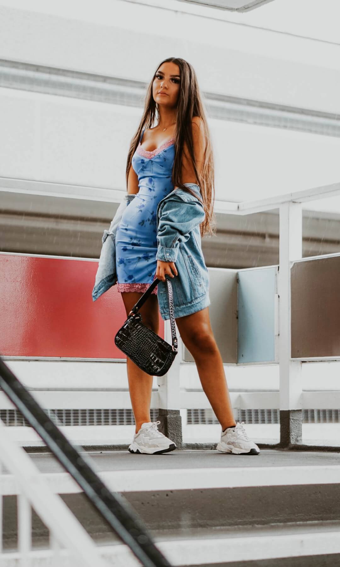 A woman stands on steps, wearing a blue dress, denim jacket, sneakers and black bag for a stylish, casual look.