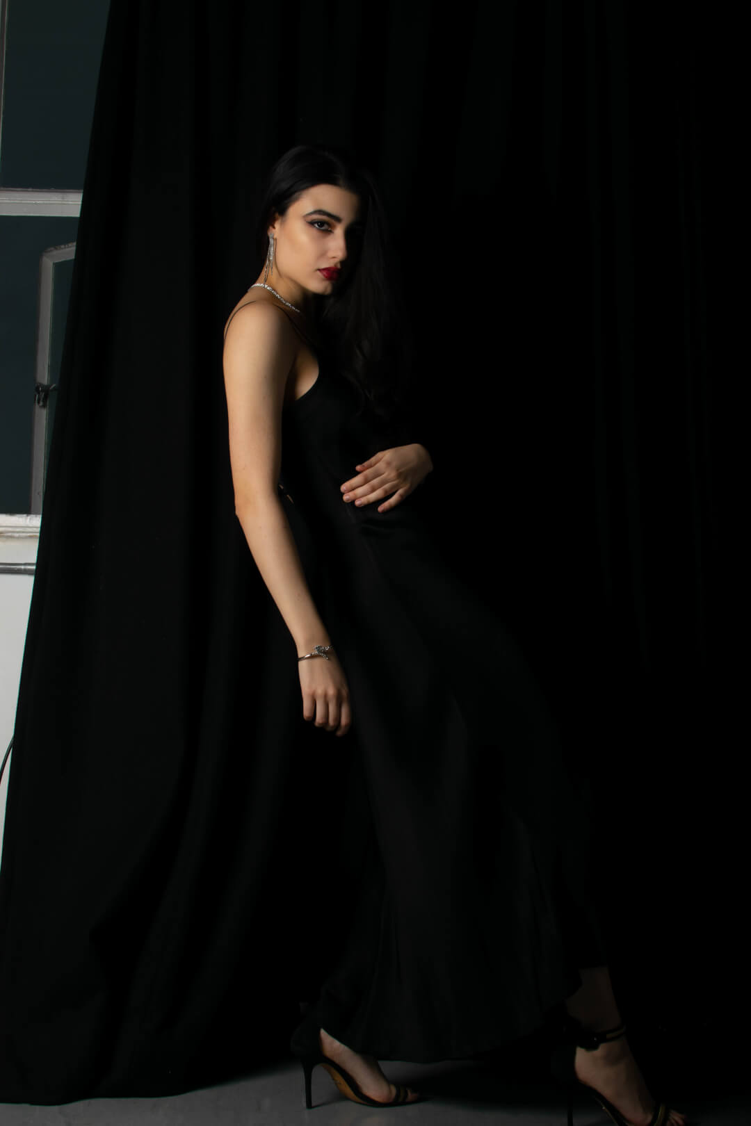 A woman in a black dress poses elegantly in a studio with jewelry and high heels.