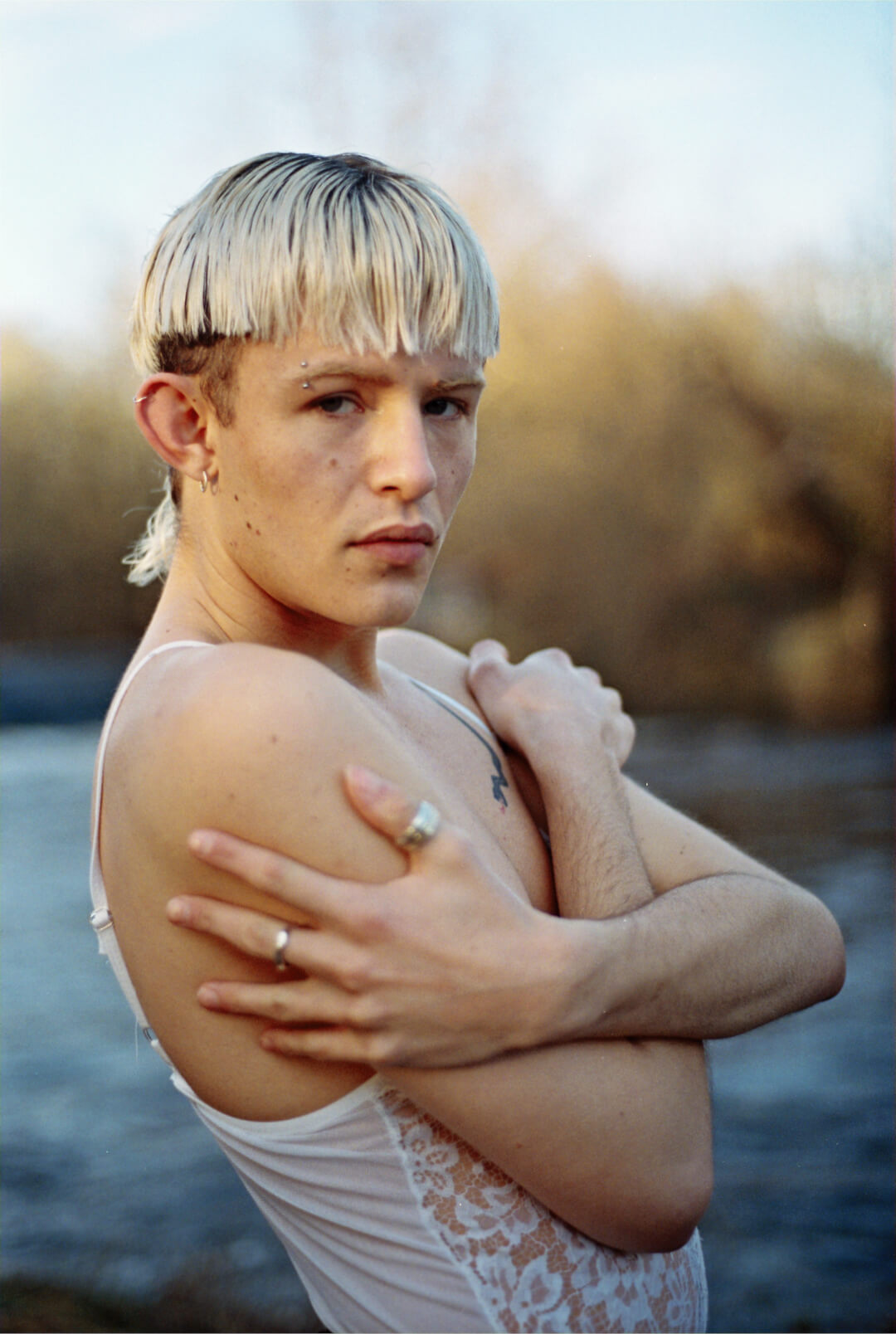 Portrait of a confident, androgynous man in a white top, posing near a river.