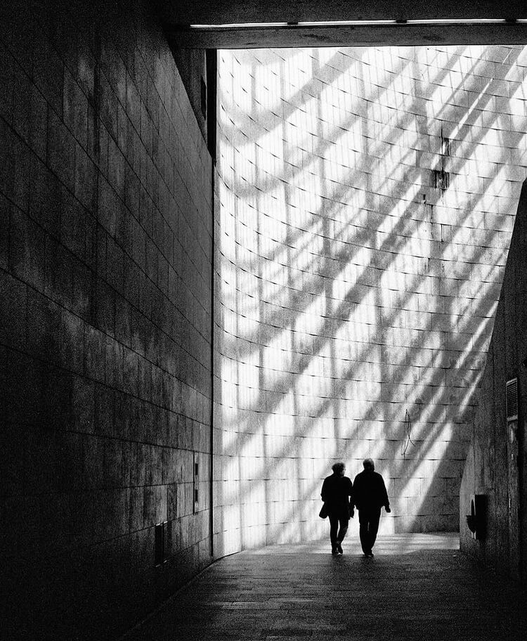 A couple walks along an alleyway, illuminated by bright light, in a monochrome image with high contrast.