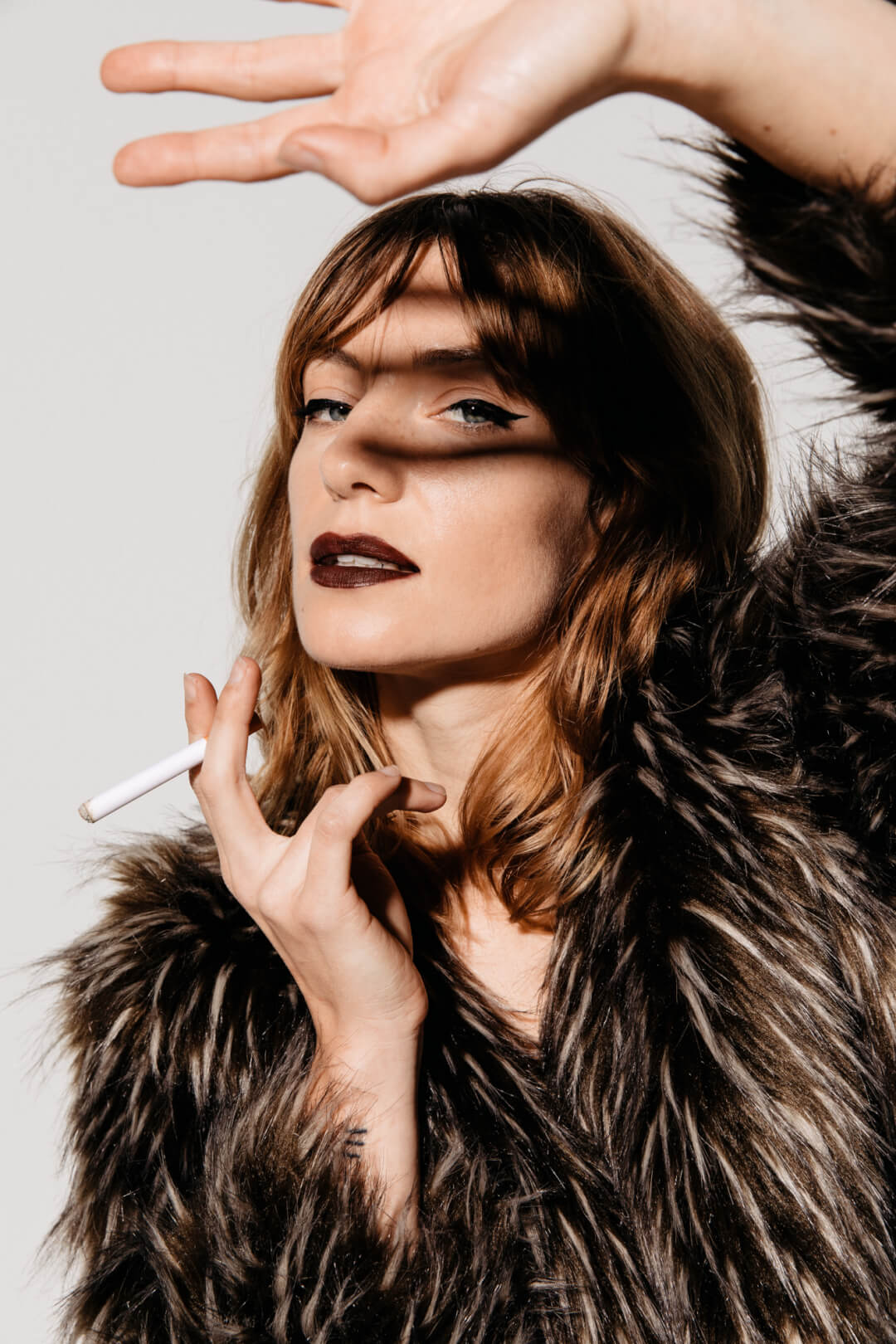 A stylish woman poses in a fur coat, cigarette in hand, with dramatic lighting for a fashion editorial.