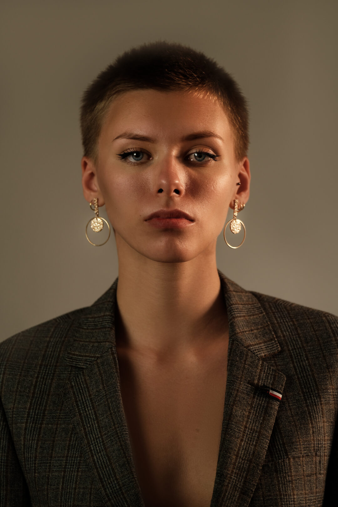 A woman with short hair wears a blazer and earrings in a studio portrait.