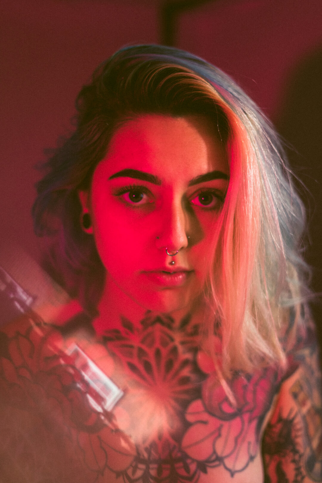 Intense portrait of a tattooed woman with striking blue and blonde hair under a red light.