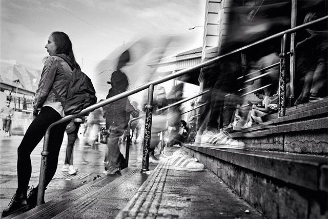 A black and white street scene captures a woman standing while others walk by with motion blur, in an urban setting.