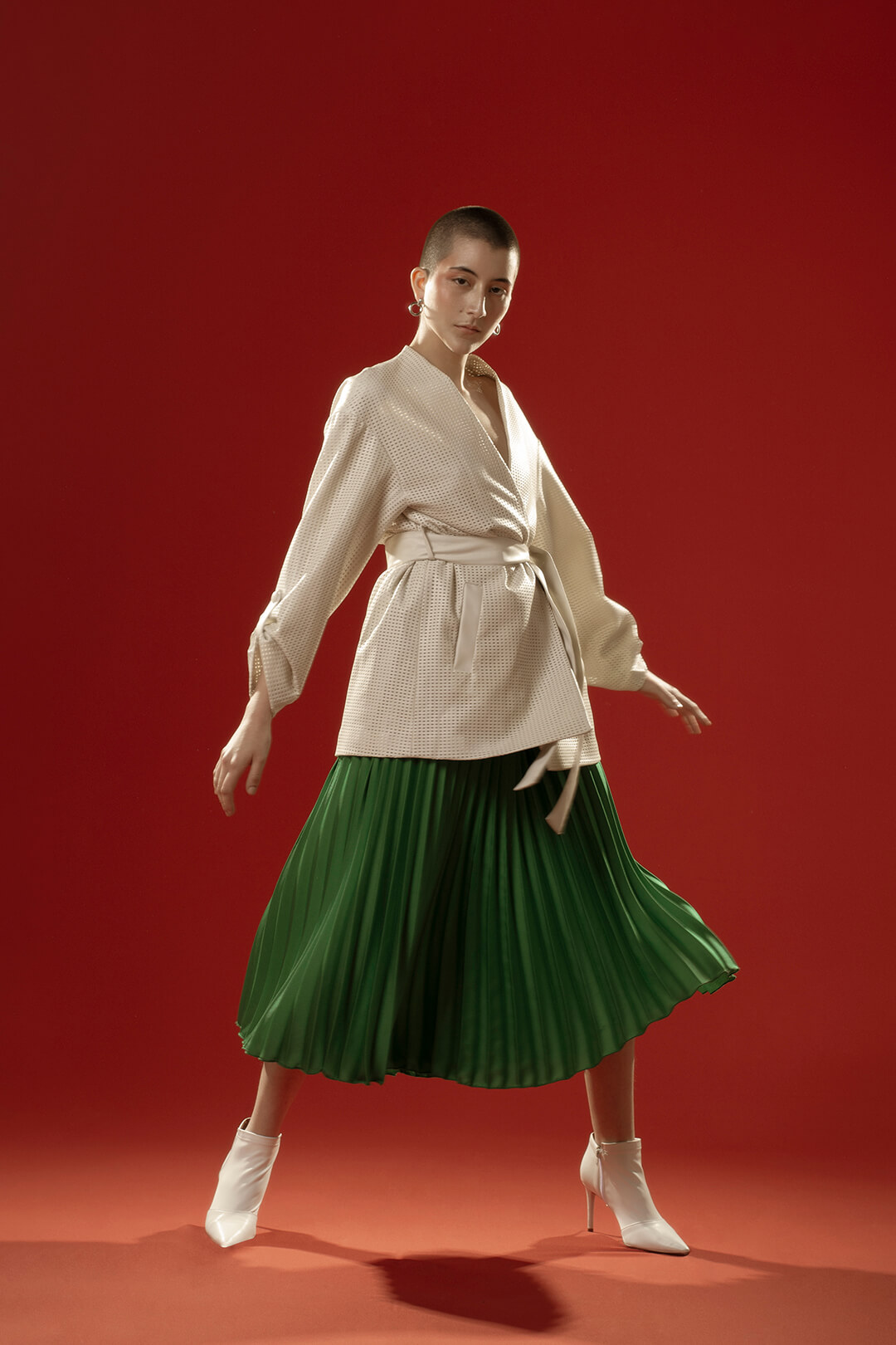 A stylish woman poses in a green skirt, blazer and white boots against a red background for a fashion editorial.