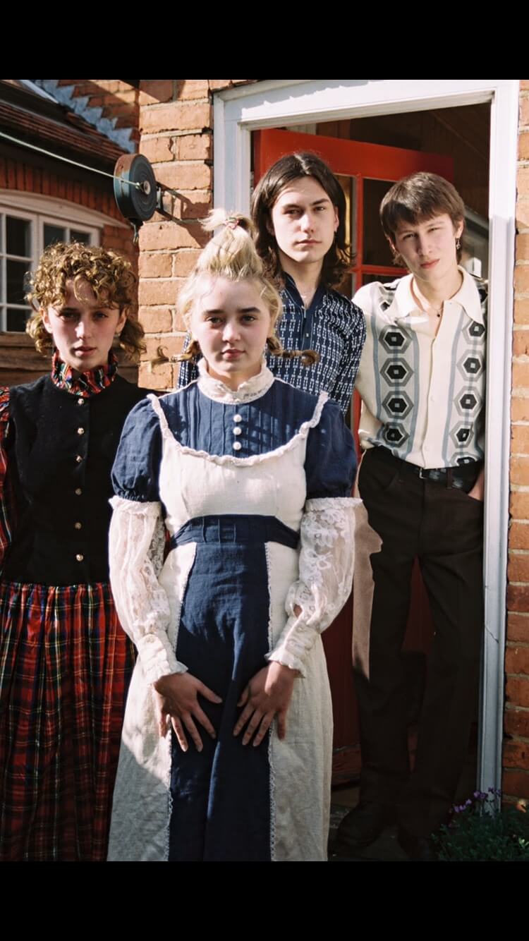 Four people in vintage clothing stand by a red brick building in soft, natural light.