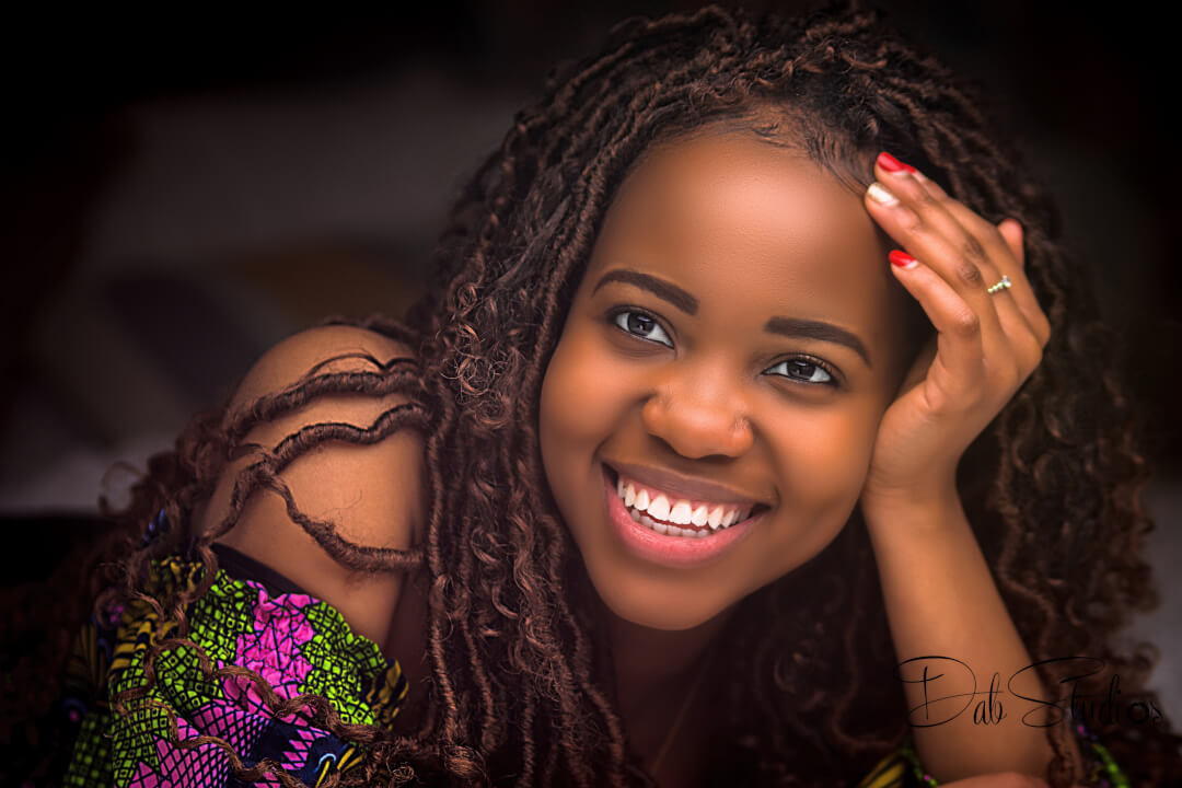 A smiling woman with dreadlocks poses in a studio with a shallow depth of field.