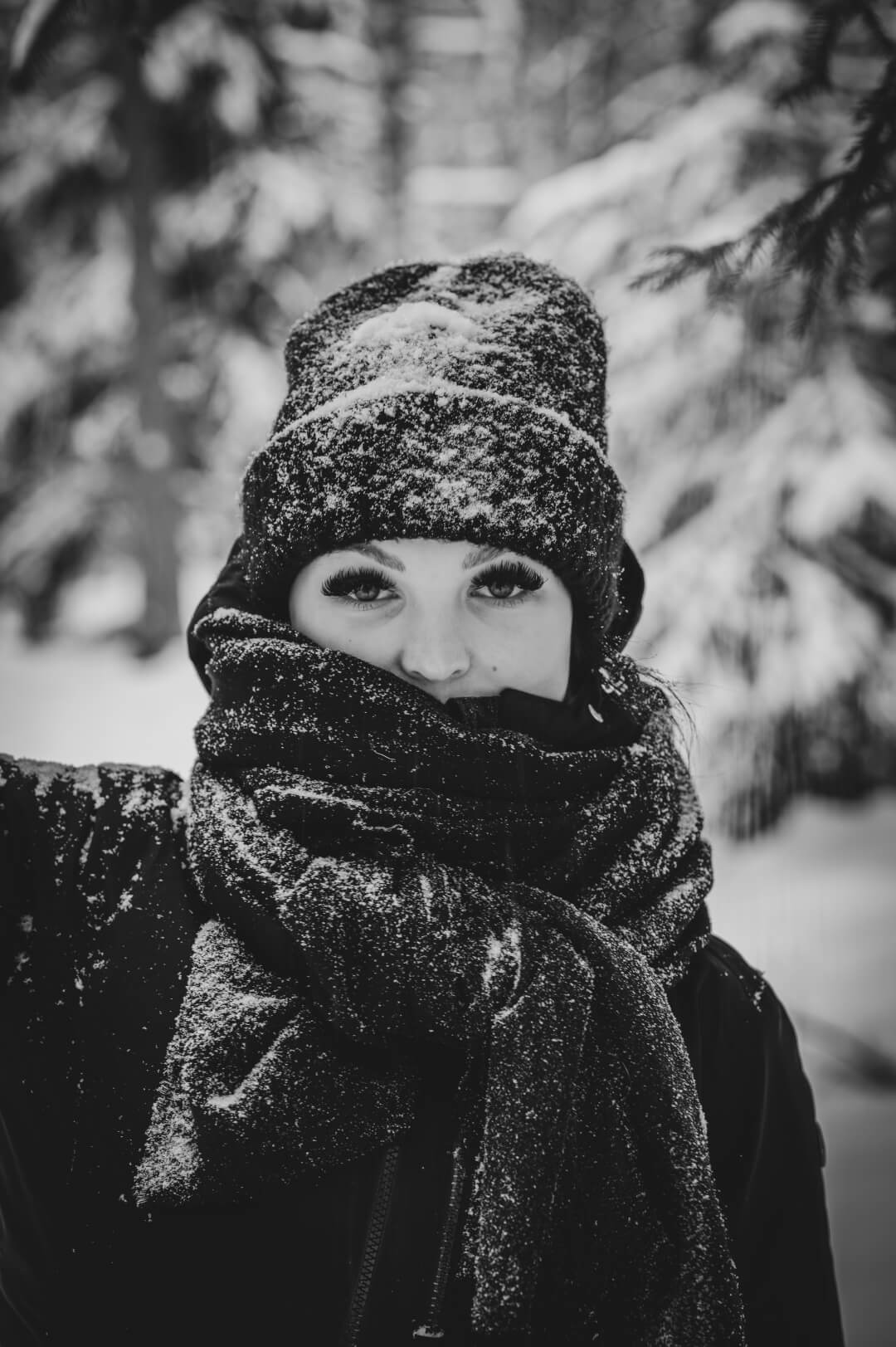 A woman bundled in a winter hat and scarf braves the snowy outdoors in a black and white portrait.