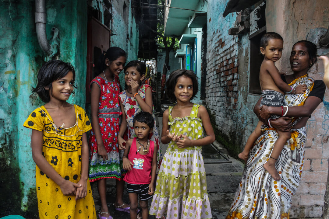 A group of children and a woman with a baby stand in a narrow alley in a vibrant community.