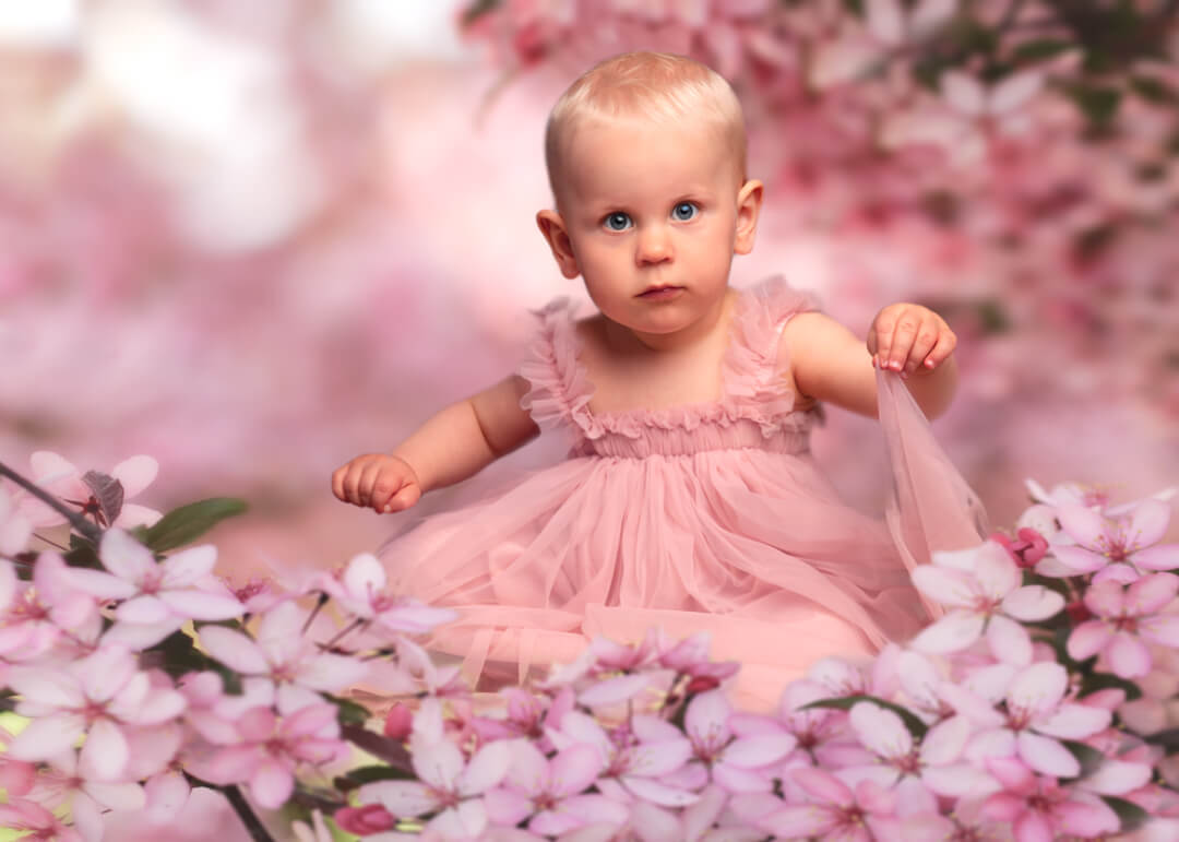 A baby girl in a pink dress sits among pink flowers in a soft, dreamy outdoor setting.