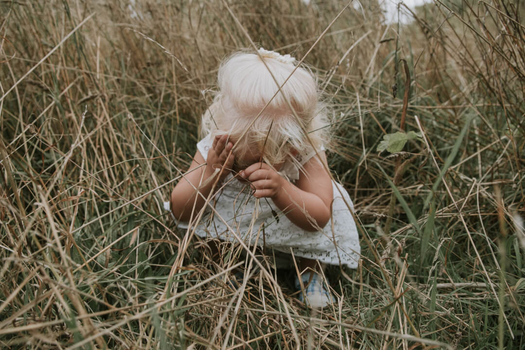 A young girl with blonde hair hides her face in a field of tall grass on a cloudy day.