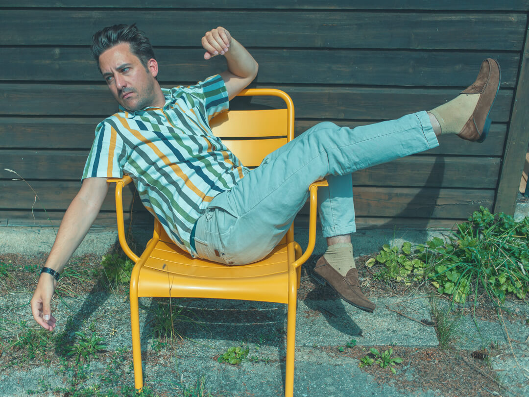 A casually dressed man relaxing on a yellow chair, set against a wood background outdoors.