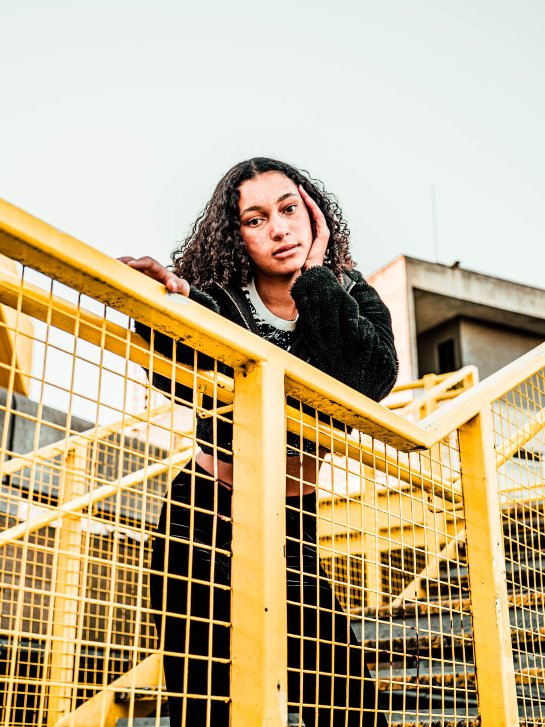 A young woman with curly hair poses on a yellow staircase in an urban setting.