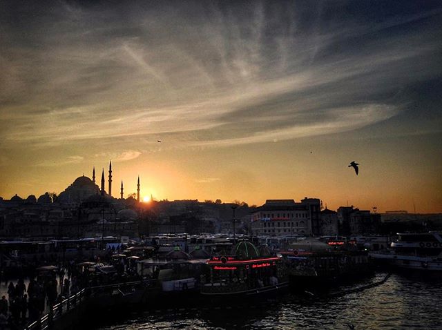 A cityscape view of Istanbul with a mosque silhouetted against a vibrant sunset sky, boats in the foreground. 