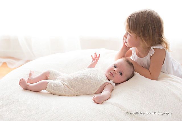 A sweet image of a baby lying next to her older sister on a soft white blanket in a brightly lit indoor setting.