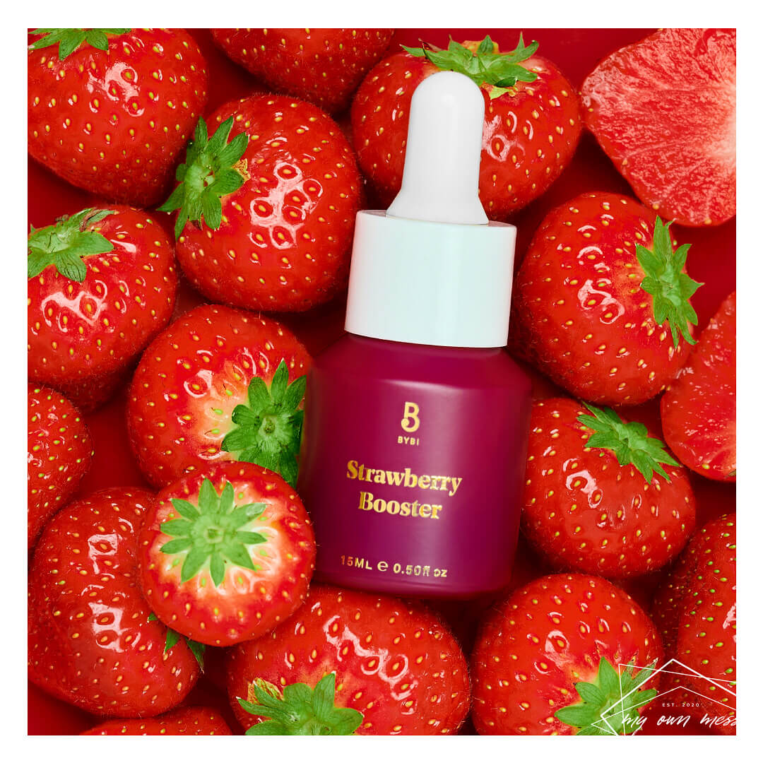 A bottle of BYBI Strawberry Booster skincare serum is surrounded by fresh strawberries.