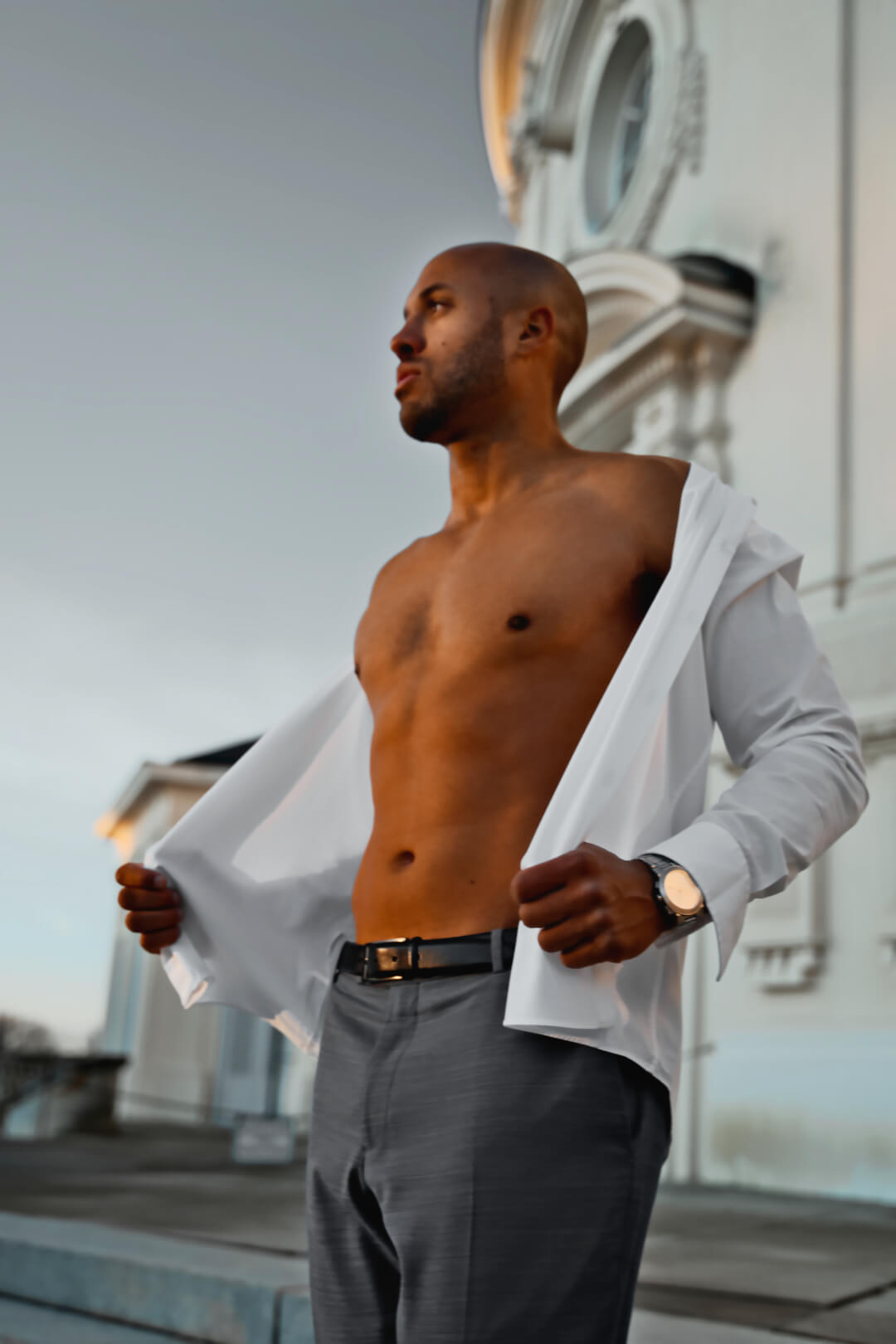 A confident bald man poses outdoors in an unbuttoned white shirt and gray pants.