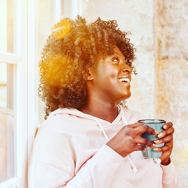 A smiling woman with curly hair enjoys a beverage in a cup by a window.