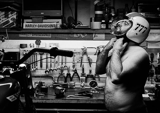 A man puts on a vintage motorcycle helmet in a garage workshop setting.