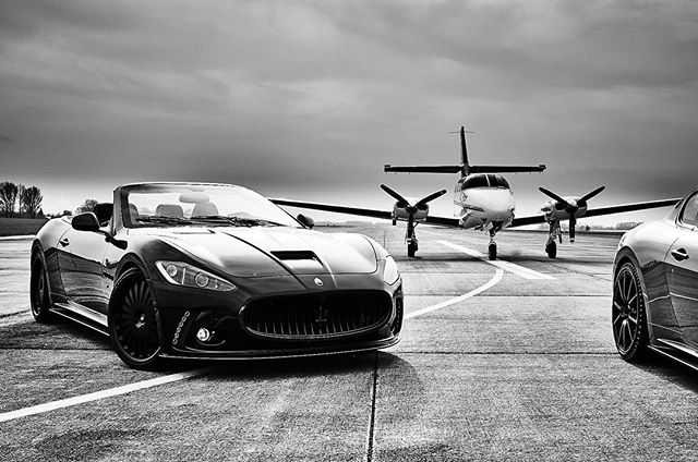 A sleek black Maserati convertible sits parked in front of a private jet on an airport runway, conveying luxury and status.