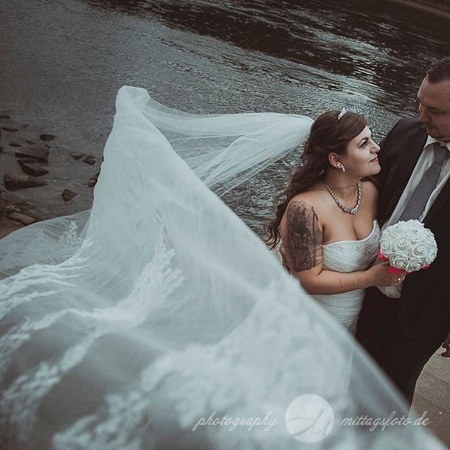 A bride and groom share an intimate moment by the river on their wedding day, with her veil flowing.