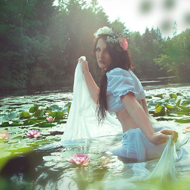 A woman with a flower crown sits in a pond filled with lilies, holding a white cloth.