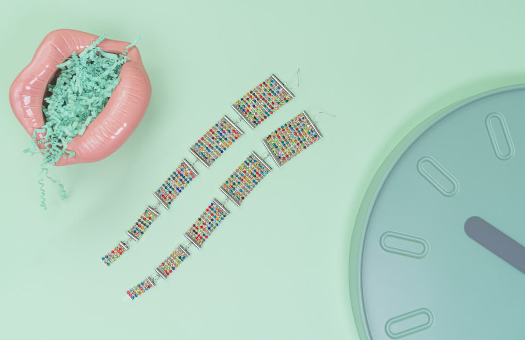 Colorful earrings are displayed with a clock and pink lips on a pastel background.