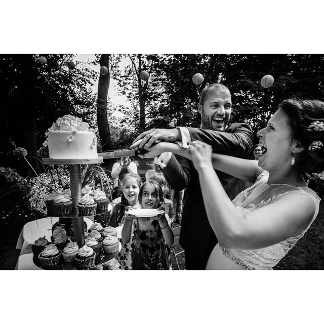 A joyful bride and groom cut their wedding cake surrounded by excited children in a candid outdoor scene.