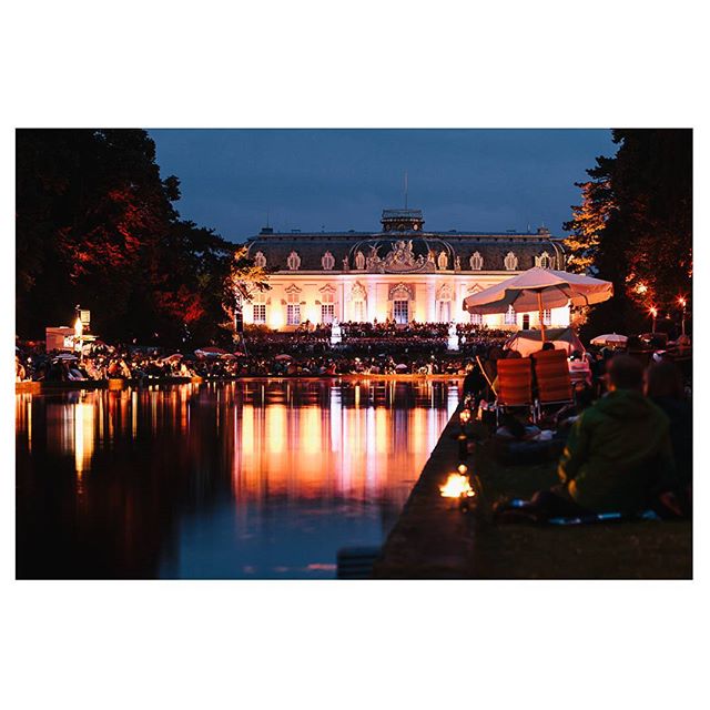 People enjoy an outdoor event at night by a lake with a building illuminated in the background, capturing a serene and lively atmosphere.