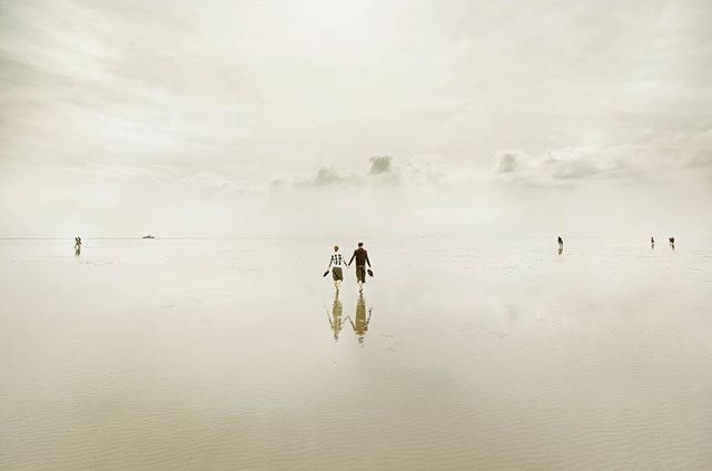 A couple walks hand-in-hand on a vast, tranquil beach under a soft, overcast sky, reflecting the calm sea.