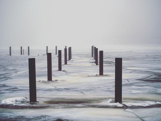A serene, foggy winter scene shows wooden posts rising from an icy lake under a hazy sky.