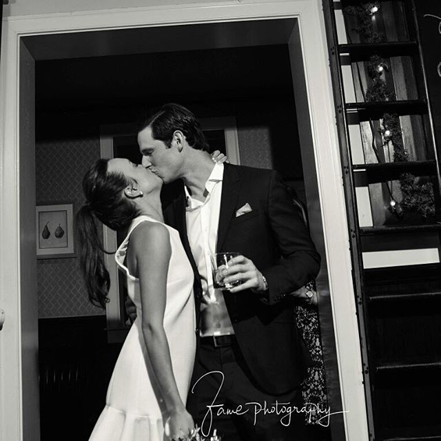 A couple in formal attire shares a tender kiss in a doorway, a timeless moment captured in black and white.