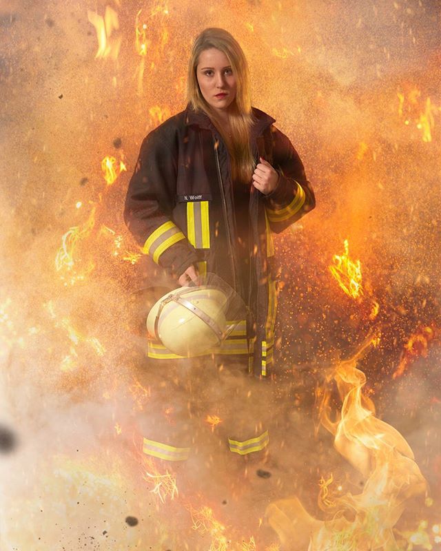 A confident woman in a firefighter's uniform stands amidst a backdrop of intense flames and smoke.