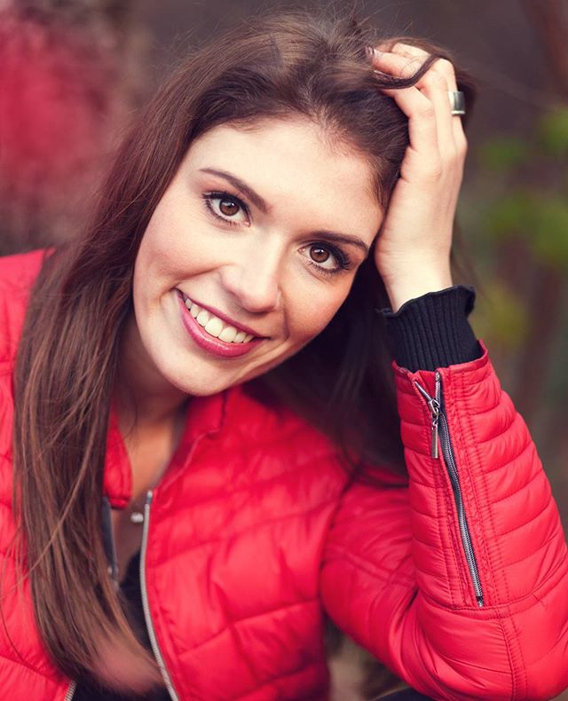 A smiling brunette woman poses in a red jacket, looking happy and cheerful.