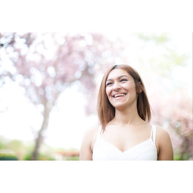 A smiling woman in a white dress enjoys a sunny day outdoors with pink blossoms in the background.