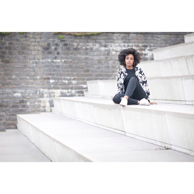 A stylish woman with curly hair sits confidently on concrete steps in an urban setting.