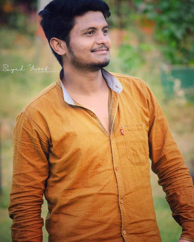 A smiling man in a brown shirt poses outdoors in a park setting.