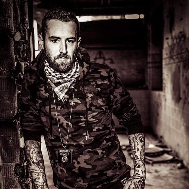 Monochrome portrait of a tattooed man in a camo hoodie leaning against a wall in an urban setting.