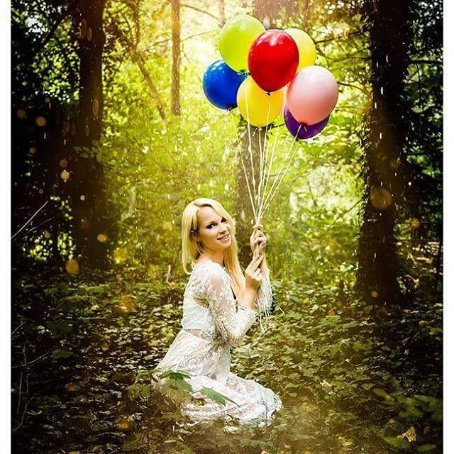 A woman kneels in a sun-drenched forest, joyfully holding a bunch of colorful balloons for a happy occasion.