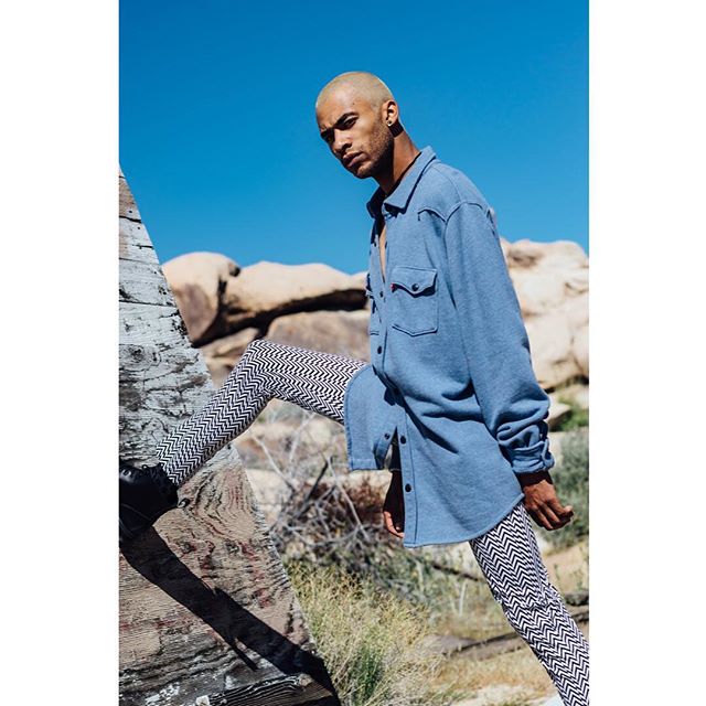 A confident male model poses in the desert wearing a blue button-down shirt and patterned leggings for a fashion shoot.
