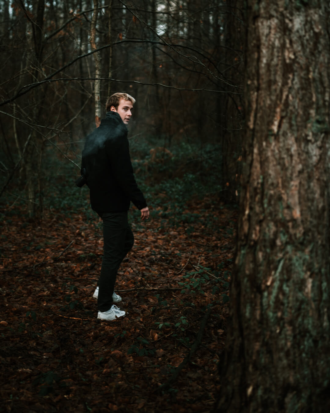 A man with blond hair walks through a dark forest, looking over his shoulder.