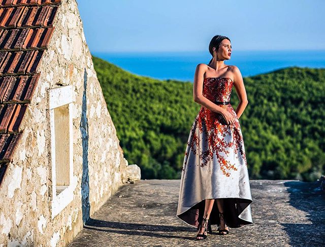 A woman in a dress stands on a rooftop with a scenic coastal and hillside backdrop.