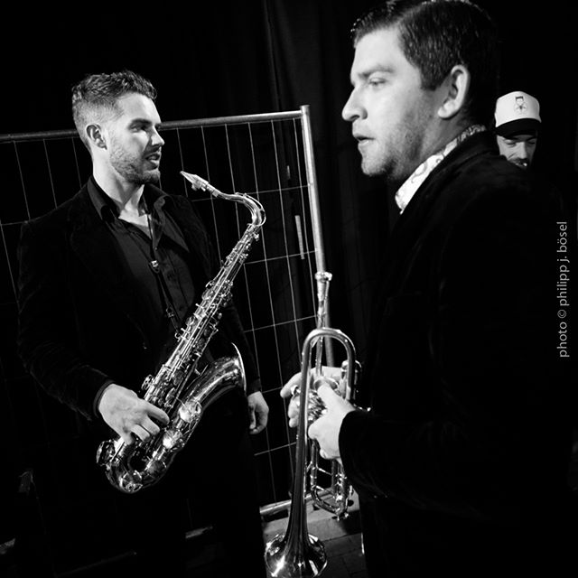 Two musicians, one playing the saxophone and the other the trumpet, performing in a dimly lit stage.