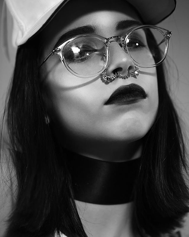 A monochrome close-up of a woman with a nose ring, glasses, black lipstick and a choker.